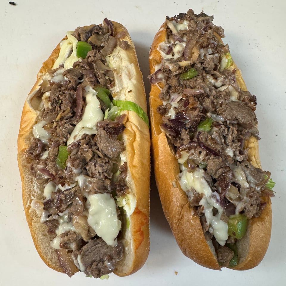 Two cheesesteak sandwiches in toasted rolls, filled with chopped beef, melted cheese, green peppers, and onions.