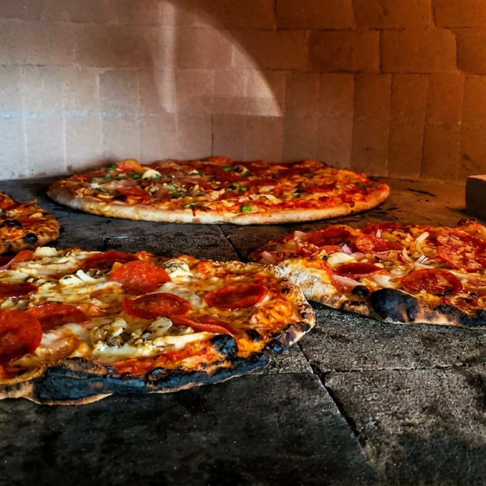 Several pepperoni pizzas bake on a stone oven floor beside a bright wood-fired flame.