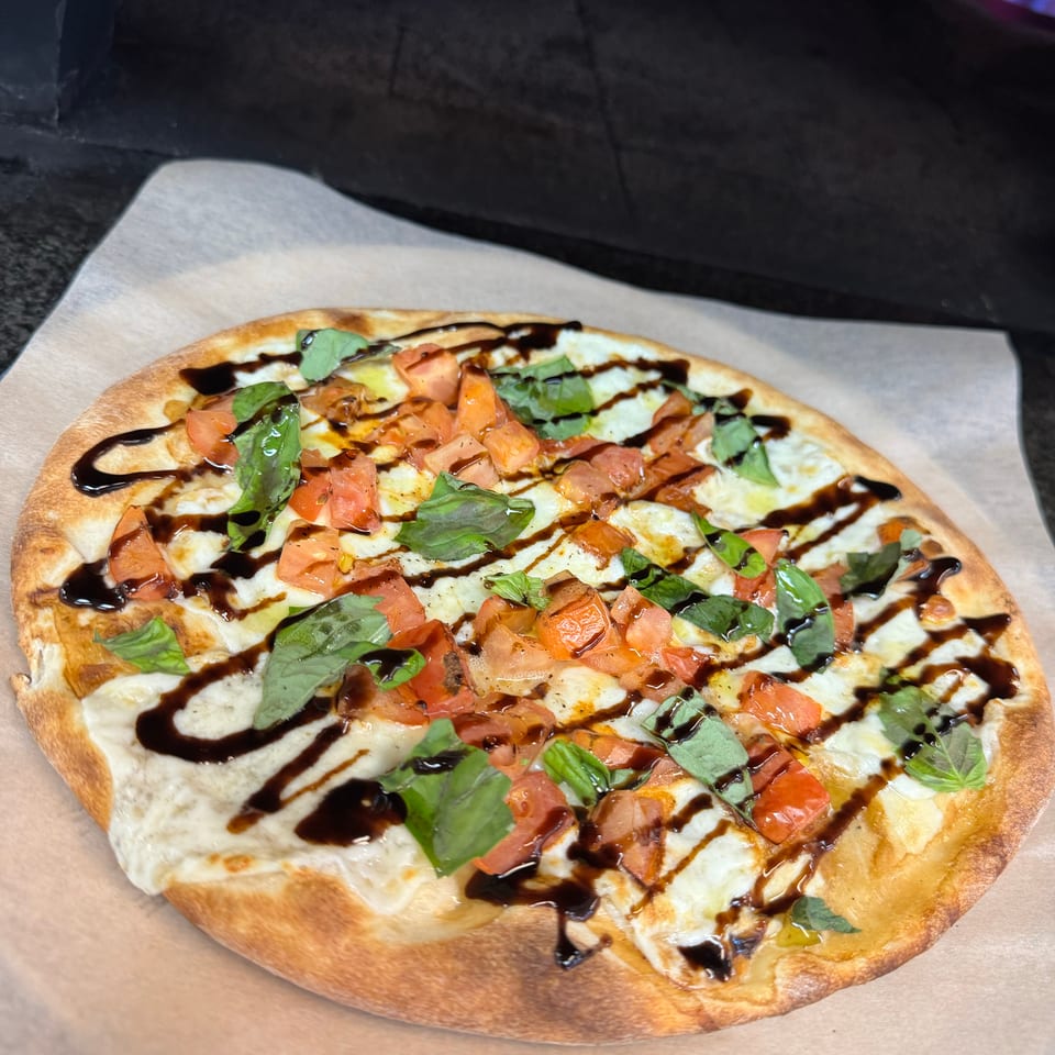 Thin-crust pizza topped with melted cheese, diced tomatoes, fresh basil, and balsamic drizzle on parchment paper.