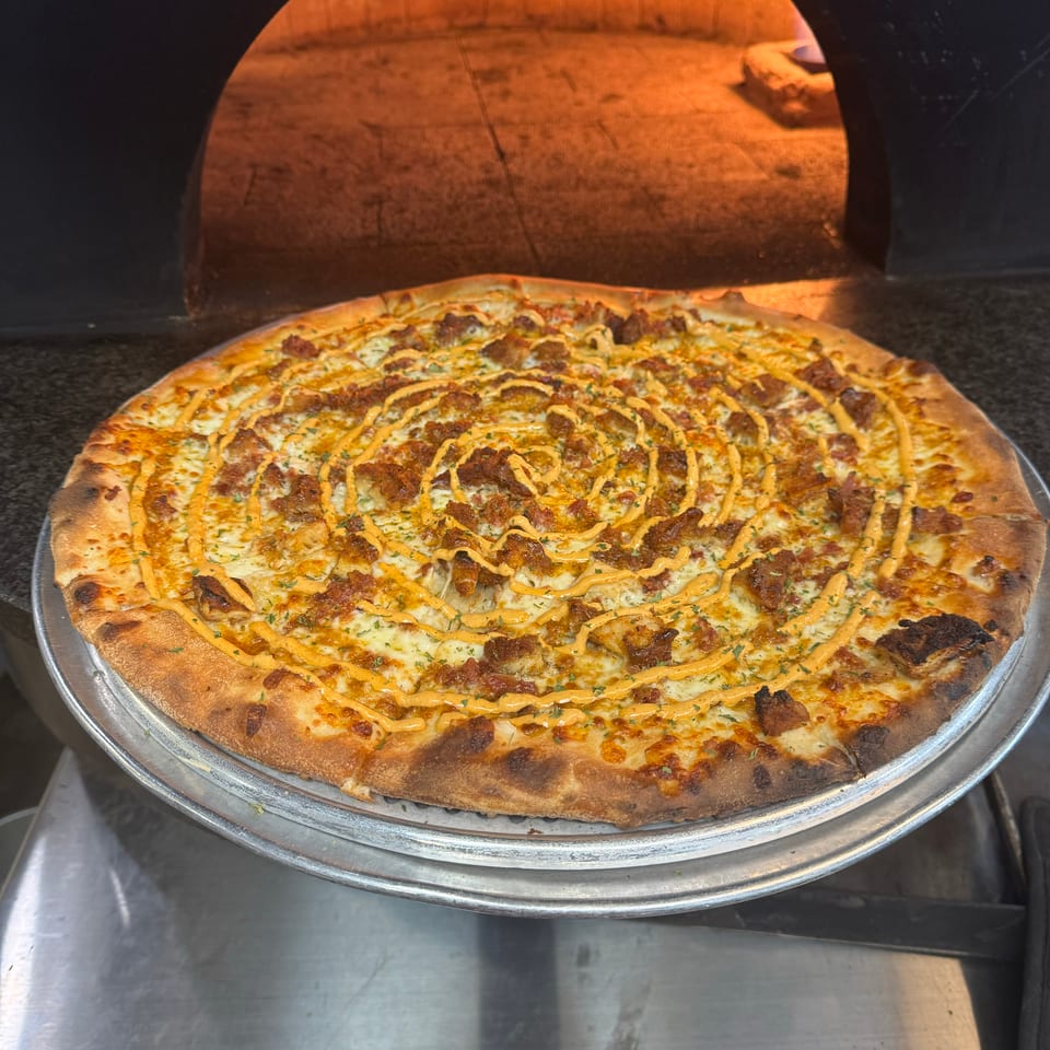 Cheesy pizza topped with browned meat, herbs, and orange sauce sits on a metal tray inside a glowing wood-fired oven.